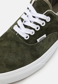 Zapatilla de ante verde, con cordones blancos, punta redonda, suela plana y una pequeña etiqueta de marca en el costado. Los detalles incluyen costuras y ojales.
