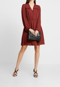 Vestido burdeos con mangas largas, escote en V y detalles de encaje. Combinado con un bolso de mano negro con grabado de cocodrilo y tacones altos negros con tiras.