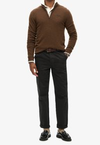 Uomo che indossa un maglione marrone con zip fino al quarto, sopra una camicia bianca con colletto, pantaloni neri con cintura marrone, calzini beige e mocassini neri con nappe.