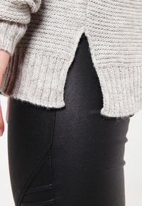 Světle šedý pletený svetr s vroubkovanými lemy, s bočními rozparky, stylizovaný přes černé legíny s koženým vzhledem a texturovaným designem.