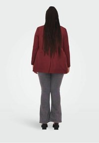 Blazer burdeos de corte relajado, combinado con jeans acampanados grises y zapatos de tacón cuadrado negros, visto desde atrás. Se puede ver el cabello largo trenzado.