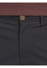 Detailní záběr na černé kalhoty s hnědým knoflíkem zapnutým na pase, spojené s hnědým tričkem zasunutým do kalhot.