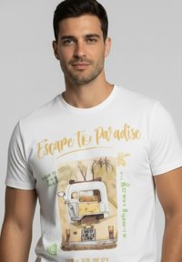 Camiseta blanca con un gráfico de una furgoneta y palmeras, texto "Escapar a Paradis  ", y acentos coloridos en verde y amarillo.