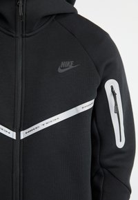 Hoodie preto Nike Tech com fecho, apresentando uma capuz, riscas prateadas e um bolso com fecho no braço esquerdo. Tecido suave e texturizado.