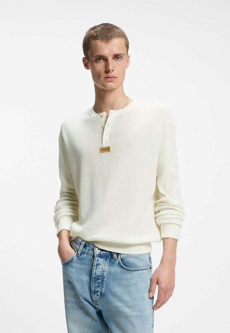 Jeune homme portant un pull henley blanc texturé à manches longues avec trois boutons et un jean bleu clair, debout avec une main dans la poche.