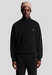 Svart fleece tröja med kvartdragkedja och logotyp, tillsammans med matchande byxor. Modellen bär en beige stickad mössa. Slät textur, avslappnad passform.