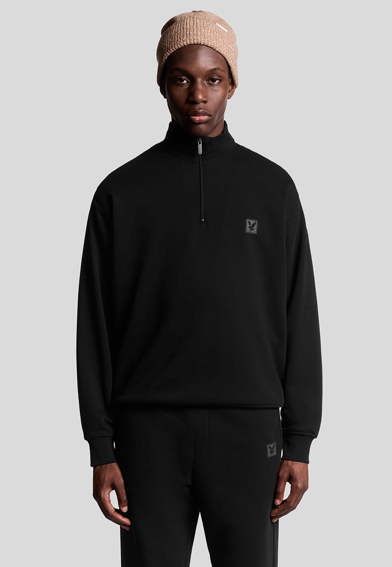 Svart fleece tröja med kvartdragkedja och logotyp, tillsammans med matchande byxor. Modellen bär en beige stickad mössa. Slät textur, avslappnad passform.