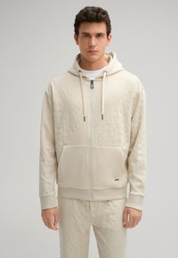 Felpa con zip color crema, dotata di un motivo testurizzato, tasca a canguro e coulisse. Polsini aderenti, pantaloni abbinati visibili, design pulito.