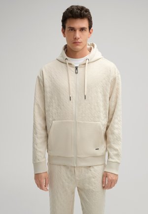 Felpa con zip color crema, dotata di un motivo testurizzato, tasca a canguro e coulisse. Polsini aderenti, pantaloni abbinati visibili, design pulito.