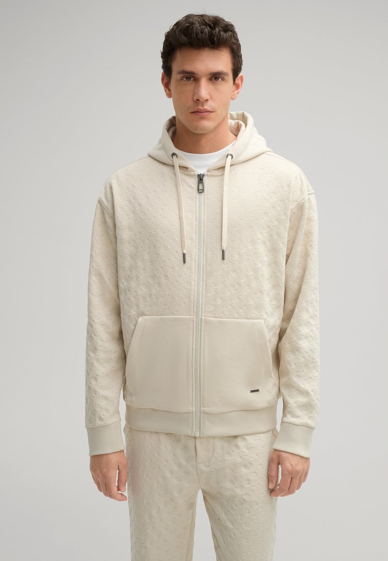 Felpa con zip color crema, dotata di un motivo testurizzato, tasca a canguro e coulisse. Polsini aderenti, pantaloni abbinati visibili, design pulito.