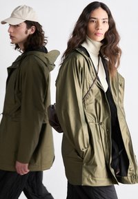 Olive green waterproof jackets with hoods, featuring drawstring waists and side pockets. Model on right carries a small brown bag.