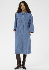 Denim srajčna obleka s ovratnikom, dvema žepoma na prsih, rokavi do komolcev, ohlapen kroj, dolžina do kolen, v svetlo modri barvi, v kombinaciji z rjavimi čevlji do kolen.