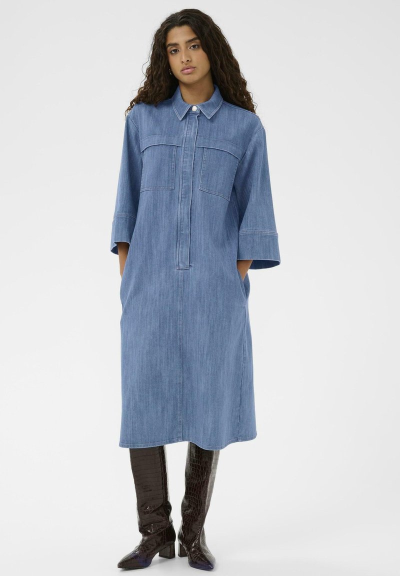 Denim srajčna obleka s ovratnikom, dvema žepoma na prsih, rokavi do komolcev, ohlapen kroj, dolžina do kolen, v svetlo modri barvi, v kombinaciji z rjavimi čevlji do kolen.