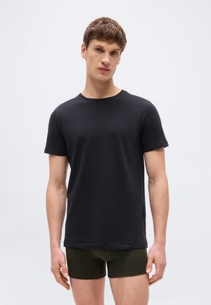 Mladý muž s krátkymi vlasmi, oblečený v jednoduchom čiernom tričku s krátkym rukávom a tmavozelených priliehavých šortkách, stojaci pred bielym pozadím.