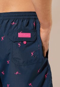 Shorts de baño azul marino con estampado de flamencos rosas, cinturilla elástica, bolsillo trasero con solapa y un parche rectangular con el logo etiquetado como "DE COSTA A COSTA."