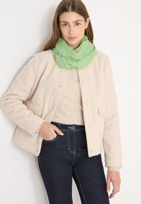 Jeune femme portant une veste polaire crème, un pull beige, un jean foncé et une écharpe vert clair autour du cou, debout devant un fond blanc.