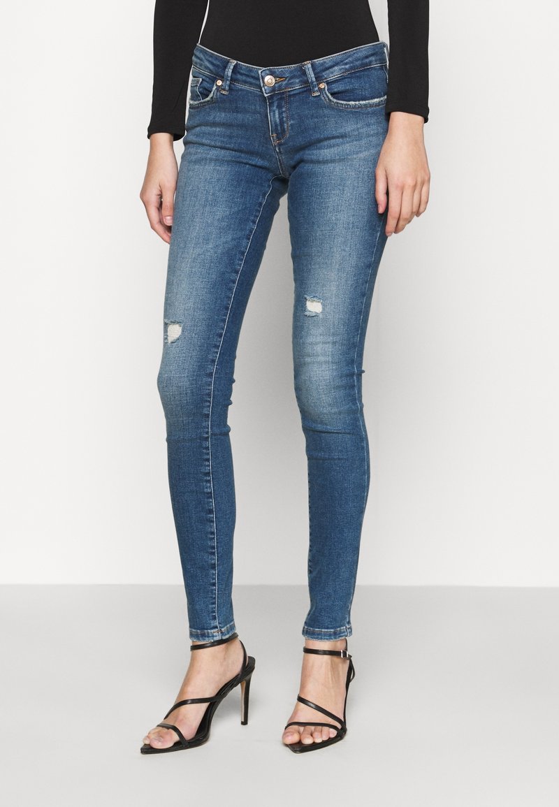 Frau trägt eng anliegende, blaue, abgenutzte Skinny-Jeans und schwarze, mit Riemchen versehene High-Heel-Sandalen, steht vor einem schlichten weißen Hintergrund.