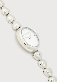 Γυναικείο ρολόι χειρός Coach σε ασημί τόνο, με οβάλ καντράν, λευκό καντράν, μινιμαλιστικούς δείκτες και βραχιόλι με συνδέσμους σε σχήμα καρδιάς.