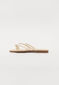 Beige flade sandaler med tre tynde stropper og en lille rund guldfarvet detalje. Glat materiale med et minimalistisk design og en brun sål.