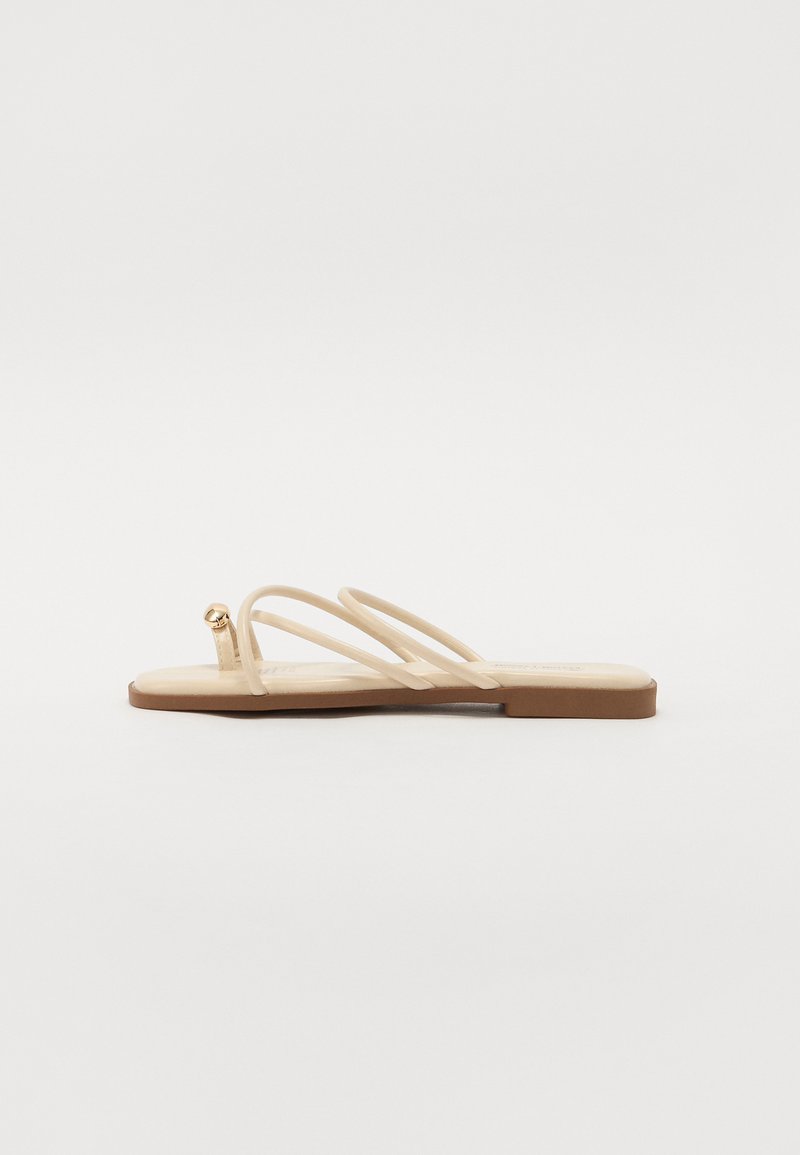 Beige flade sandaler med tre tynde stropper og en lille rund guldfarvet detalje. Glat materiale med et minimalistisk design og en brun sål.