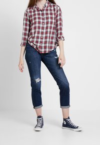 Camicia a quadri rossa e blu, maniche arrotolate; jeans scuri strappati blu; sneaker alte navy con accenti bianchi.
