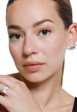 Silberne, blattförmige Ohrringe und passender Ring; strukturiertes Design mit kunstvollen Details. Das Modell präsentiert die Schmuckstücke vor einem weißen Hintergrund.