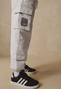 Pantaloni cargo gri deschis cu buzunar aplicat, decorat cu un design grafic negru. Asortați cu adidași negri cu dungi albe și șosete albe.