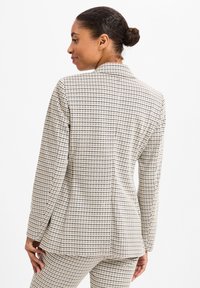 Maßgeschneiderter Blazer in Creme mit einem schwarz-roten Karomuster. Verfügt über einen Reverskragen, strukturierte Schultern und einen einzelnen Rückenschlitz.