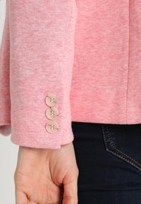 Manica di giacca a maglia rosa con tre bottoni rotondi di colore chiaro e una sottile texture. Jeans in denim neri visibili sotto.