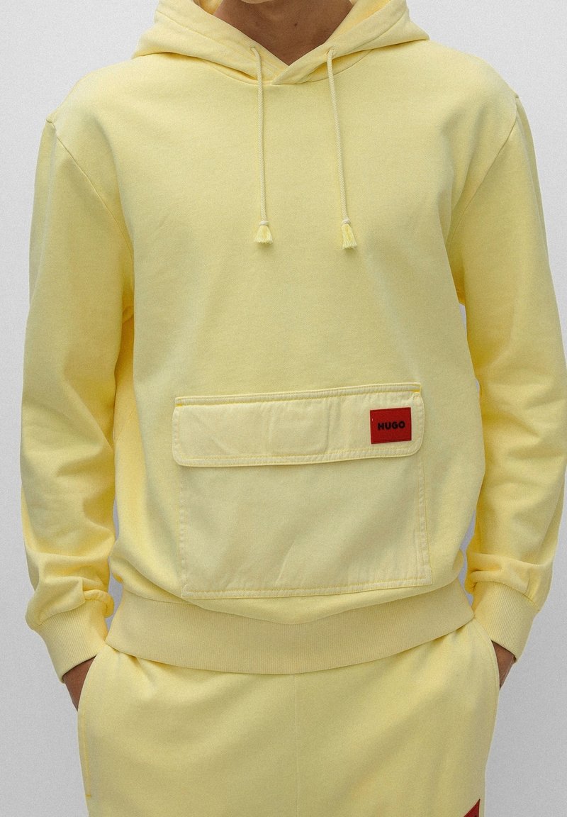Personne portant un sweat à capuche jaune pâle avec une poche à rabat à l'avant ornée d'une étiquette rouge "HUGO", assorti à un pantalon jaune.