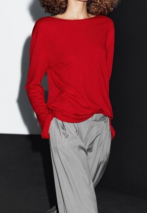 Personne aux cheveux bouclés portant un haut rouge à manches longues ample et un pantalon plissé gris taille haute, les mains dans les poches.