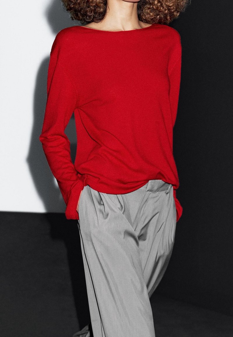 Personne aux cheveux bouclés portant un haut rouge à manches longues ample et un pantalon plissé gris taille haute, les mains dans les poches.