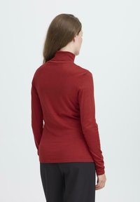 Sarkanais augstais kakls džemperis izgatavots no mīkstas auduma. Pieguļošs dizains ar ribotām piedurknēm un apakšmalu. Nēsāts kopā ar melnām biksēm, veidojot vienkāršu, plūstošu izskatu.