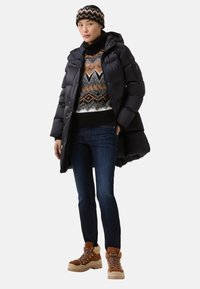 Černá péřová bunda s kapucí, barevný vzorovaný svetr, tmavě modré džíny a hnědé turistické boty s kožíškem a šněrováním.