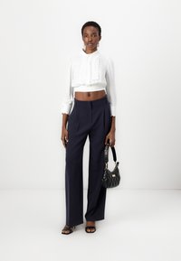 Blusa bianca cropped con dettagli scallop e maniche lunghe, abbinata a pantaloni neri a palazzo. Tiene in mano una borsa a tracolla nera trapuntata.