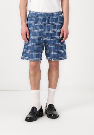 Mand iført blå teksturerede ternede shorts, hvide crew-strømper, sorte læder loafers og en hvid t-shirt, står mod hvid baggrund.