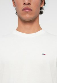 Ung mand iført hvid T-shirt med rund hals og et lille rektangulært logo i rød, hvid og blå på venstre bryst, let ansigtshår og en øredobb.