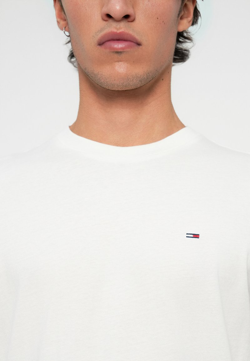 Joven con camiseta blanca de cuello redondo con un pequeño logo rectangular rojo, blanco y azul en el lado izquierdo del pecho, ligero vello facial y una pendiente.