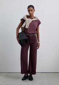 Mujer vestida con un top sin mangas color burdeos y pantalones de pierna ancha, suéter beige drapeado sobre los hombros, sosteniendo una chaqueta de cuero negra, con mocasines negros.