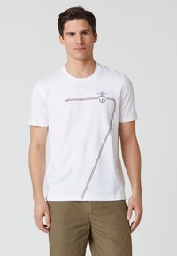 T-shirt bianco a maniche corte in cotone, con un design rosso e marrone e un'emblema sulla parte superiore sinistra. Classico scollo rotondo.