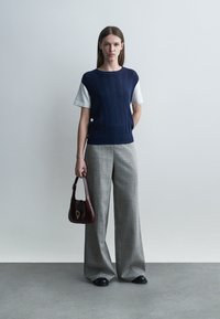 Kvinne står mot en ensfarget vegg iført marineblå ermeløs genser over hvit skjorte, grå vide bukser, svarte sko, og holder en mørkebrun håndveske.