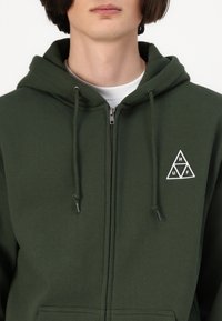 Grønn zip-up hettejakke laget av mykt stoff, med et trekantet hvitt logo på venstre bryst, justerbare snorer og kengurulommer.