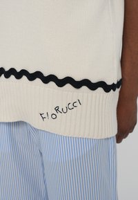 Maglione lavorato a maglia di colore crema con bordi ondulati neri all'orlo e logo "Fiorucci" ricamato; indossato sopra pantaloni a righe blu.