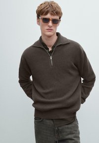 Brauner Rippenstrickpullover mit Reißverschlusskragen, getragen über einem dunklen Hemd, kombiniert mit hellbrauner rechteckiger Sonnenbrille.