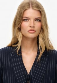 Marineblauwe pinstripe blazer met een slank, op maat gemaakt ontwerp. Voorzien van afgeronde revers, een enkele knoopsluiting en een gestructureerde stof.
