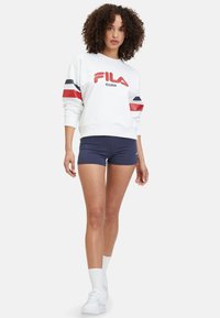 Witte FILA sweatshirt met rode en marineblauwe strepen op de mouwen, met een gedurfde logo. Gecombineerd met marineblauwe shorts en witte sneakers.