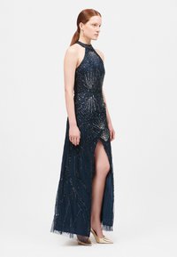 Abito da sera in blu navy con dettagli in perline, scollo all'americana, spacco laterale e sovrapposizione in tessuto trasparente. Completato da una siluetta aderente e tacchi dorati.