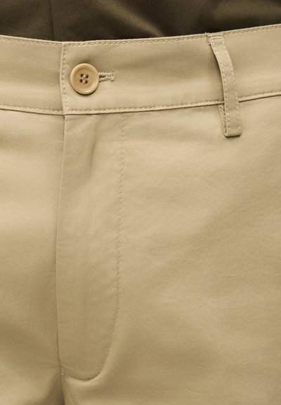 Gros plan sur la ceinture d’un pantalon beige avec fermeture à bouton et passants pour ceinture, porté avec un haut vert olive foncé.