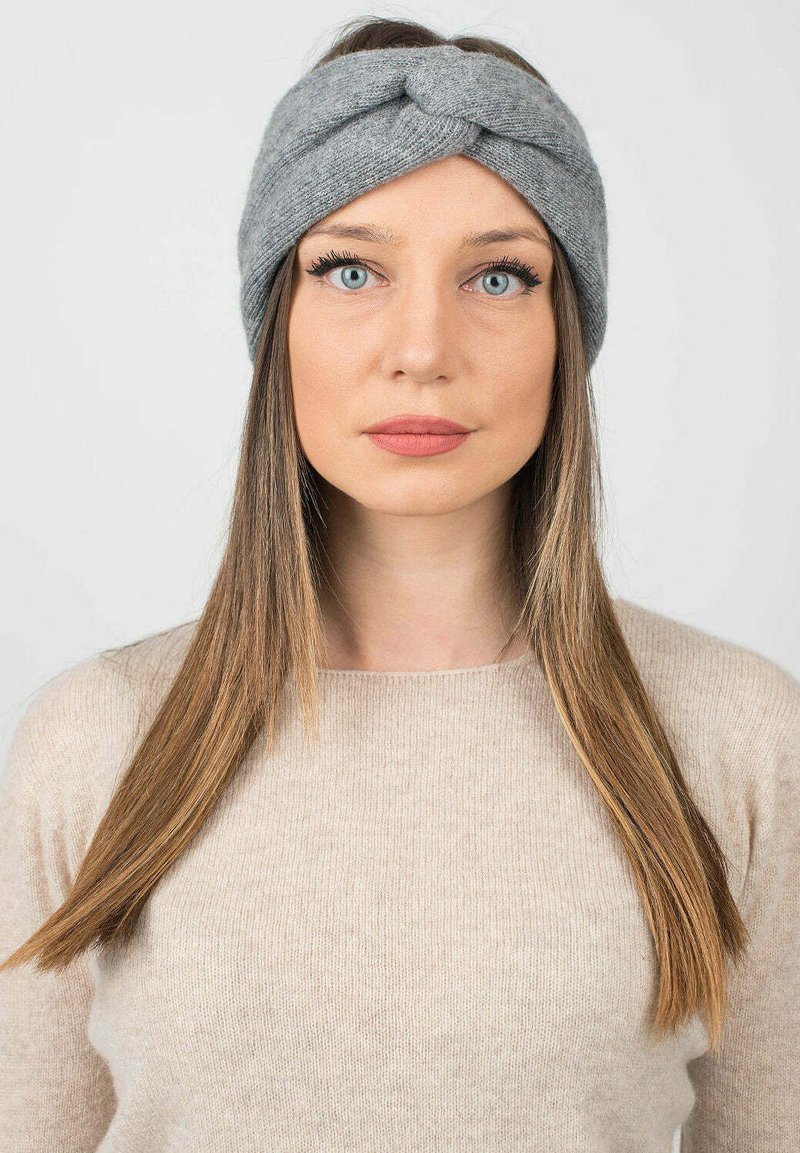 Fascia in maglia grigia con design attorcigliato nella parte anteriore, indossata su capelli lunghi e lisci, abbinata a un maglione di colore chiaro.