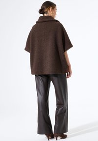 Brauner strukturierter Poncho mit hohem Kragen, weiten Ärmeln und offener Silhouette, kombiniert mit dunkelbraunen Lederhosen und Absatzschuhen.